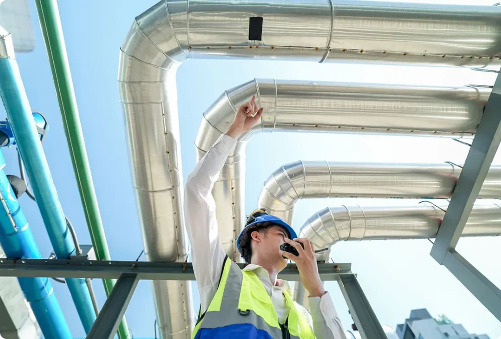 Person with reflective vest and hard hat talking on walkie-talkie with silver tubes above